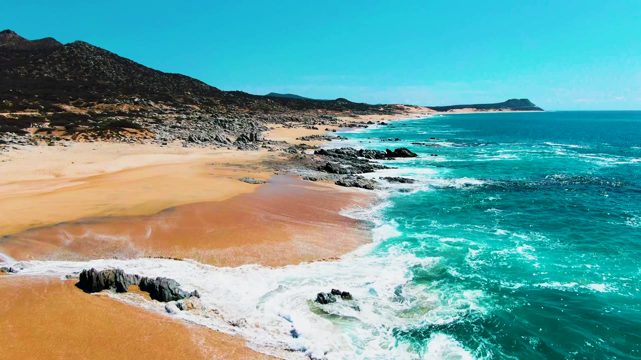Drone-style aerial view at Punta Buzzards, Baja California Sur, where waves break around dark rocks beside broad golden sand, with open ocean to the right and no visible people.