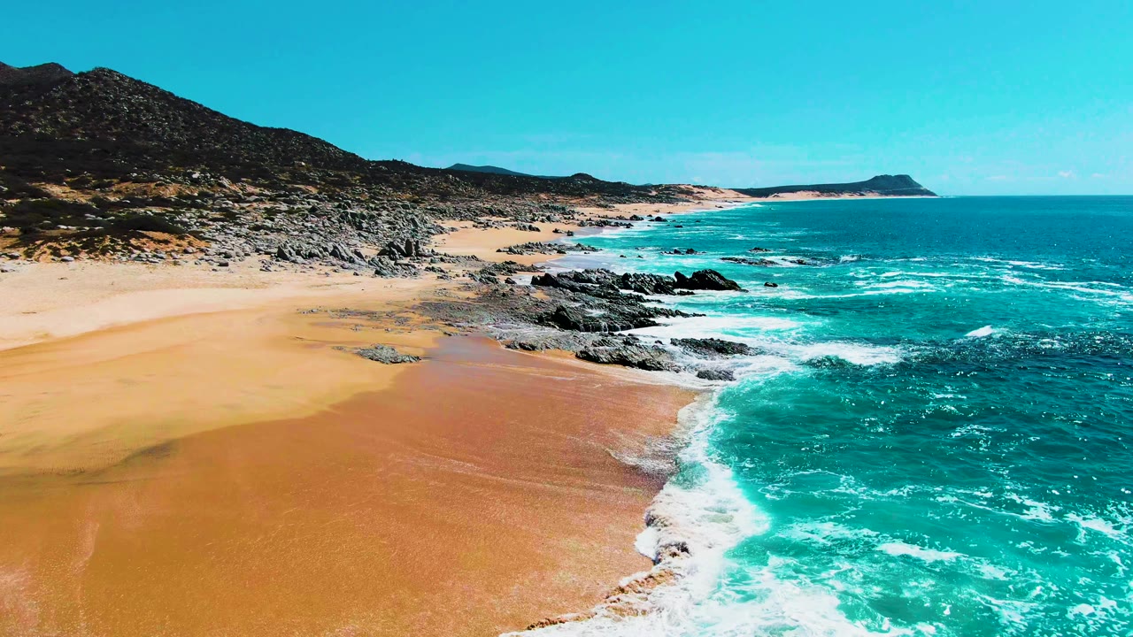 Wide aerial frame at Punta Buzzards, Baja California Sur, showing a larger stretch of rocky shoreline, orange-gold wet sand, turquoise sea, distant coast, and no visible people.