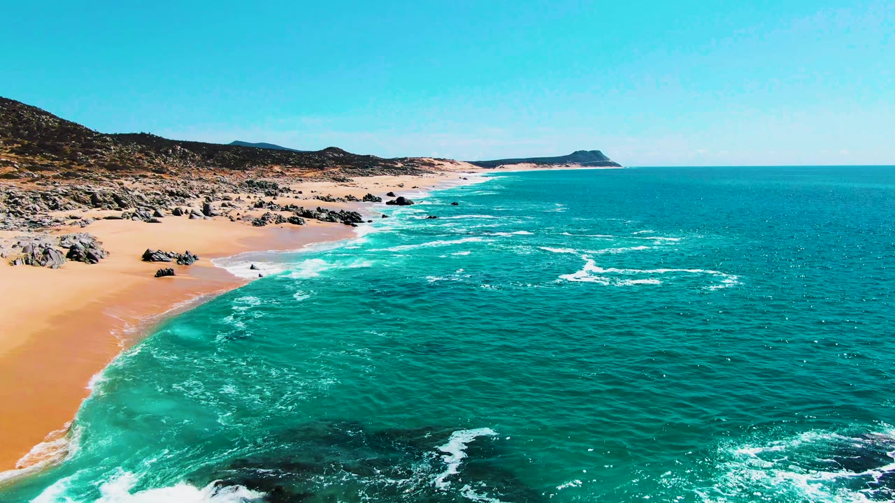 Aerial view of turquoise ocean breaking onto a sandy beach with rocks and low hills.