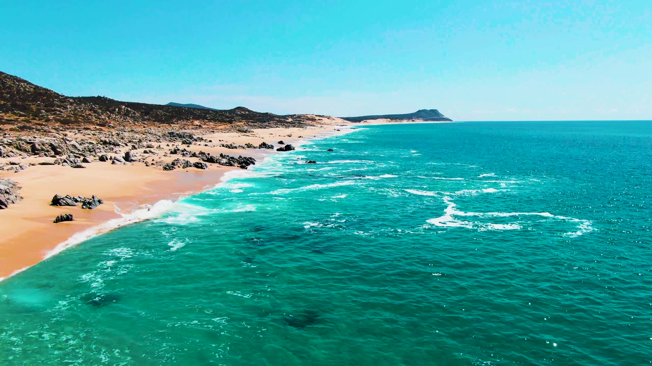 At one second, the aerial view sits farther along the surf line at Buzzards Beach, with white foam spreading across shallow turquoise water, rocks on the sand, and no visible people.