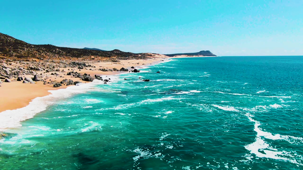 At three seconds, the drone view shows surf pushing up the sand at Buzzards Beach, with dark rocks clustered along the left shoreline, clear turquoise water below, and no visible people.
