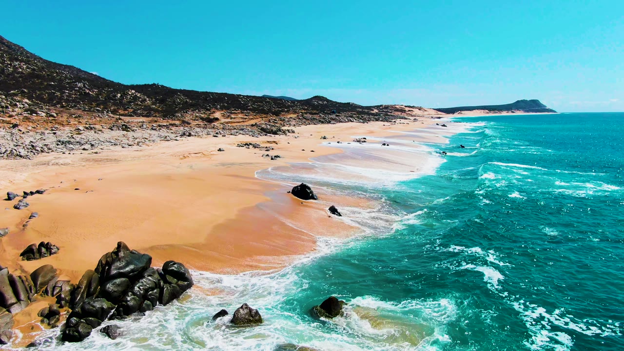 Aerial view at Punta Buzzards, Baja California Sur showing turquoise surf, golden sand, dark foreground rocks, low hills, a distant headland, and no visible people.