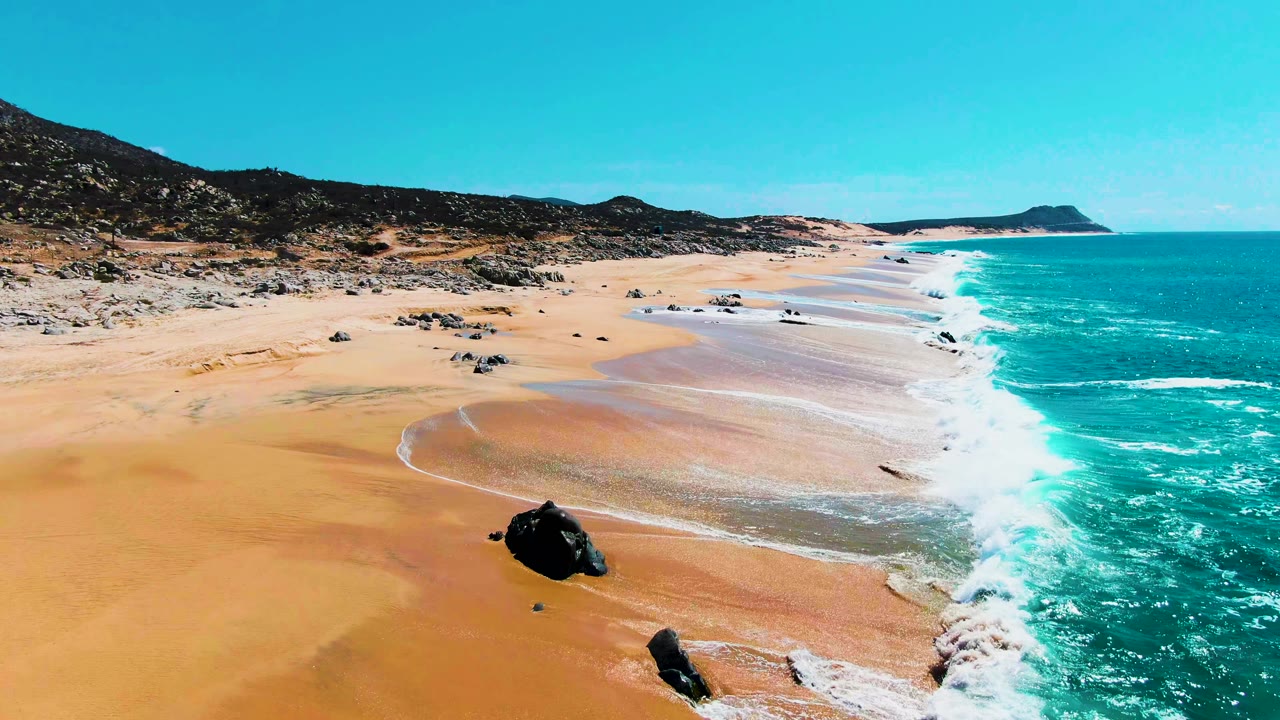 Aerial coastline frame at Punta Buzzards, Baja California Sur showing surf sliding up a broad sandy beach, scattered dark rocks, turquoise sea, and no visible people.