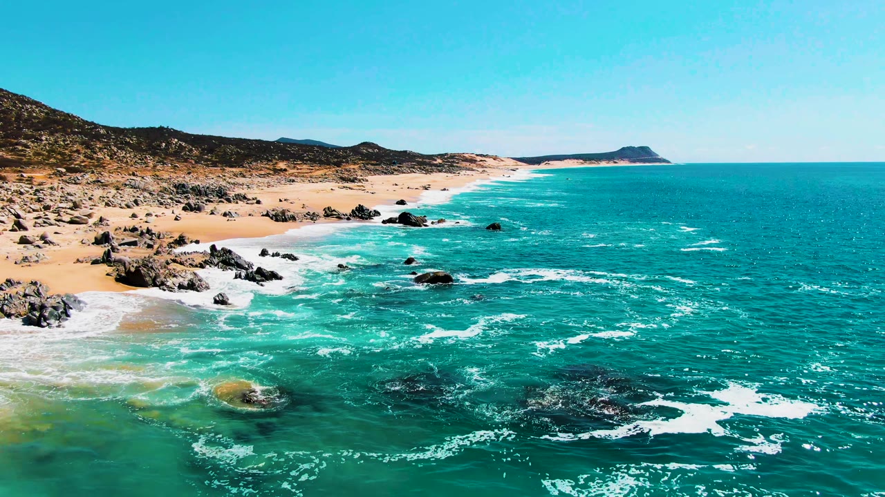 Aerial view over Buzzards Beach in Baja California Sur, Mexico, with turquoise waves breaking around dark rocks beside tan sand and no visible people.