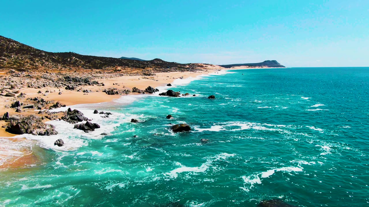 Buzzards Beach shoreline seen from above, with whitewater spreading through blue-green water around offshore rocks and no visible people.