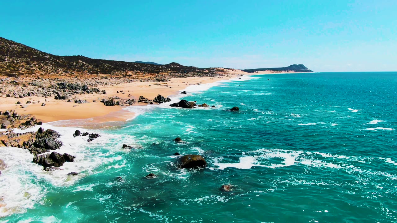 Wide aerial frame at Buzzards Beach showing waves curling toward the sand, dark rocks in the surf, a distant headland, and no visible people.