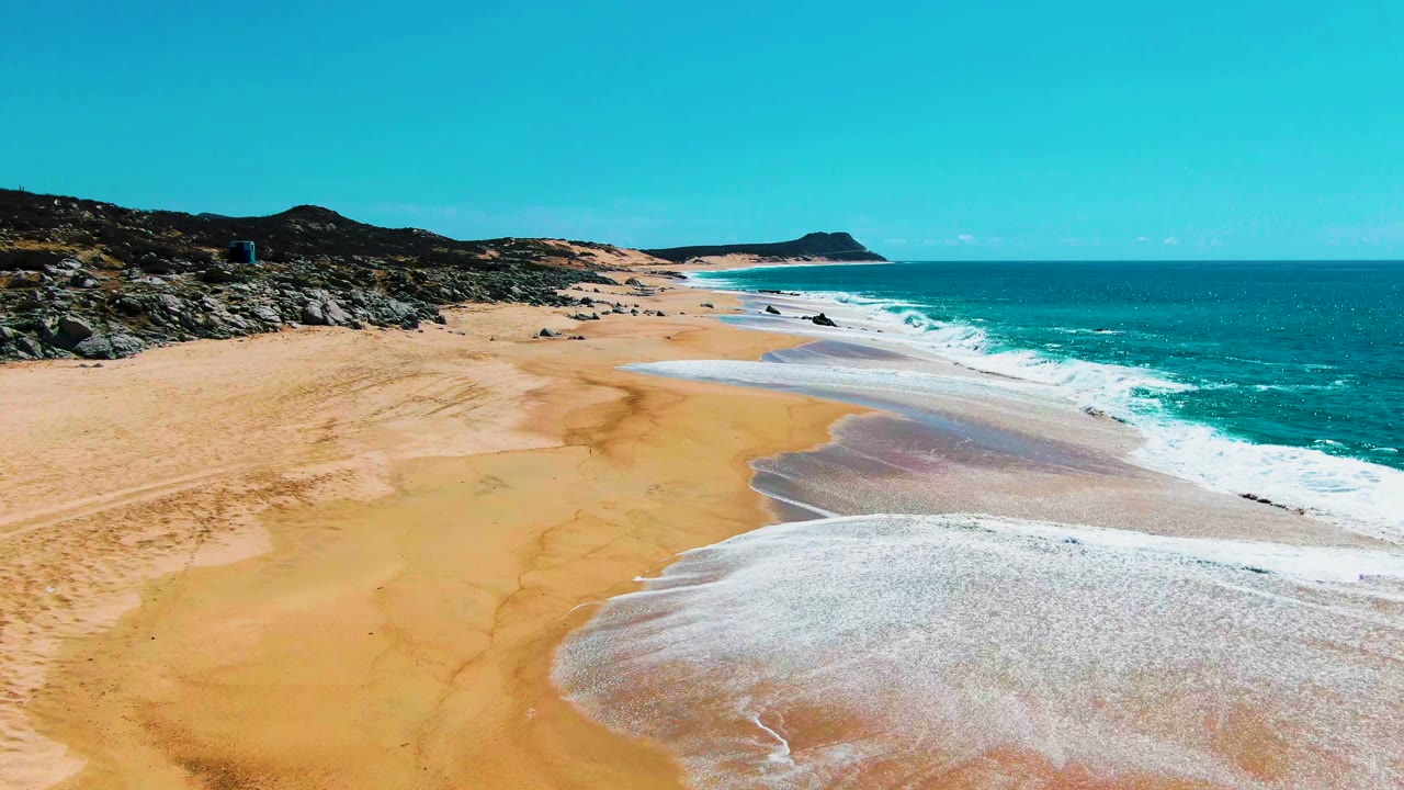 Aerial view of turquoise surf washing onto a wide sandy beach with rocks at left.