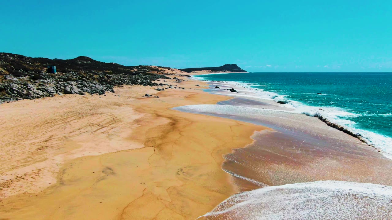 Aerial view at Punta Buzzards with a broad sheet of foam over the lower beach, darker wet sand tracing the waterline, rocks along the left shore, and no visible people.