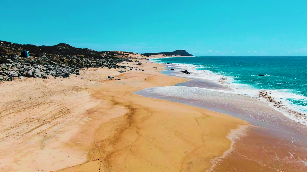 Punta Buzzards aerial frame showing the surf line pulled farther along the right shore, a broad open sand area in the center, rocky terrain to the left, and no visible people.