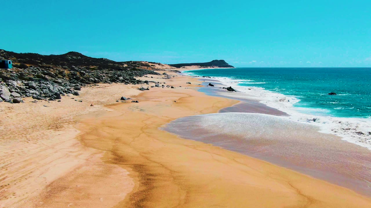 At Punta Buzzards, the aerial composition shows wet sand bands in the foreground, white surf along the right edge, darker rocks on the left slope, and no visible people.