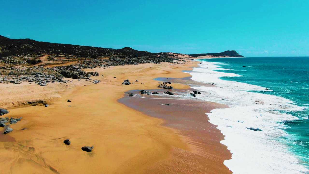 Aerial view of Punta Buzzards beach in Baja California Sur with golden sand, scattered rocks, turquoise water, white surf, low hills, and no visible people.