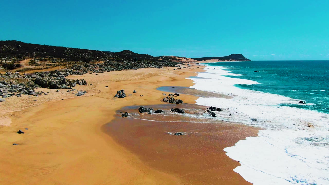 Wide aerial frame over Punta Buzzards showing wet brown sand near the waterline, dark rocks in the surf zone, turquoise ocean, white foam, and no visible people.