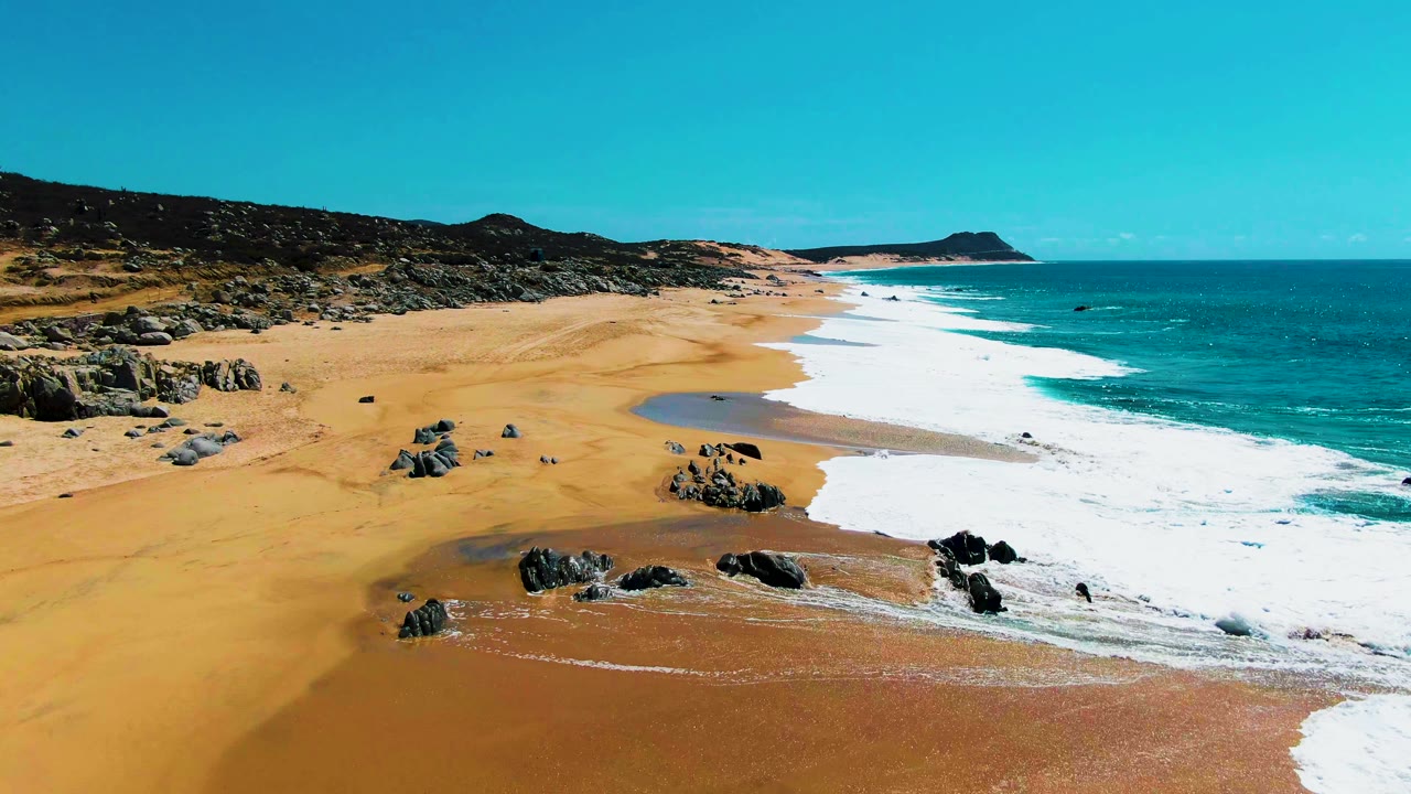 Aerial view at Punta Buzzards with white surf curling around rocks on the sand, open turquoise water to the right, low hills behind the beach, and no visible people.