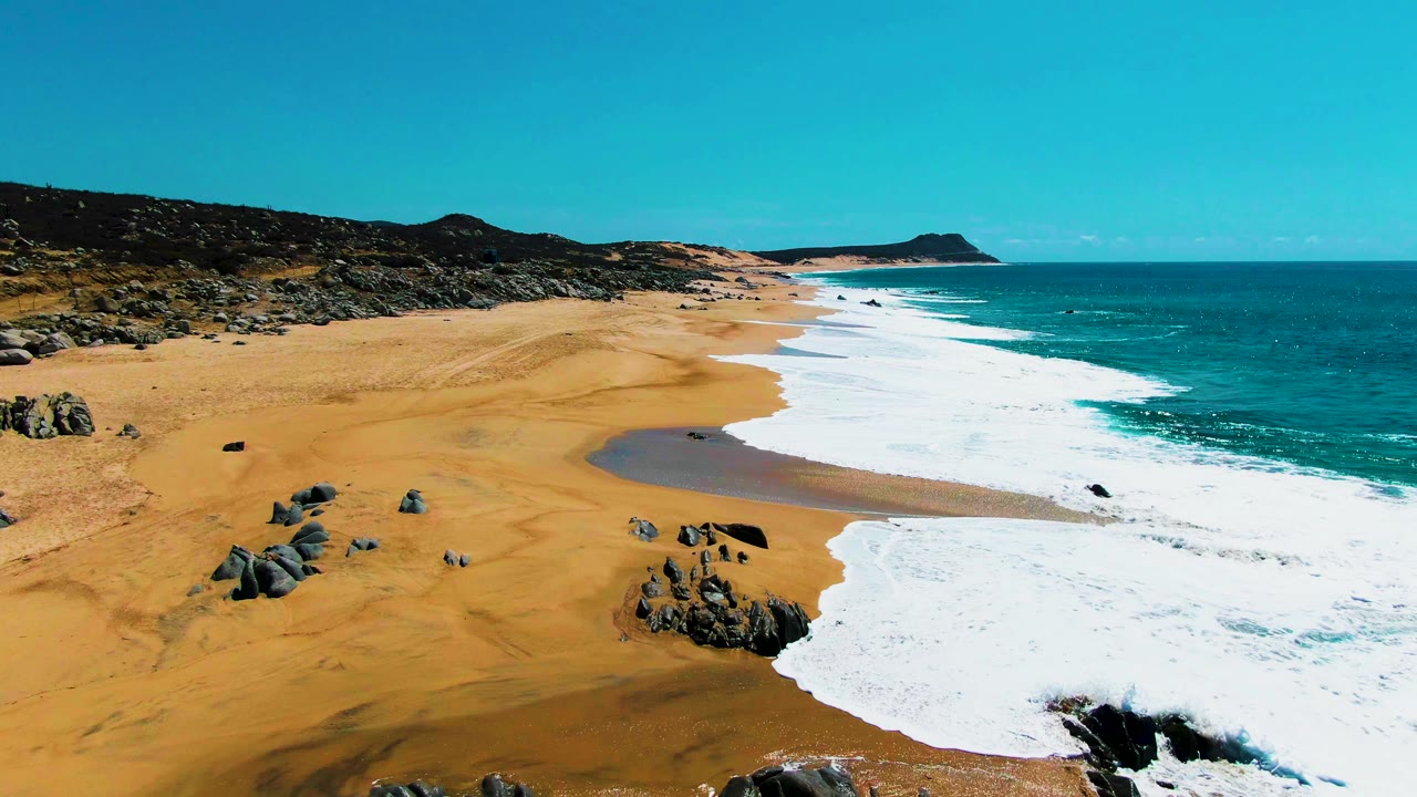 Oblique aerial frame of Punta Buzzards with rocks in the lower beach area, broad golden sand, white foam pushing up the shoreline, turquoise water, and no visible people.