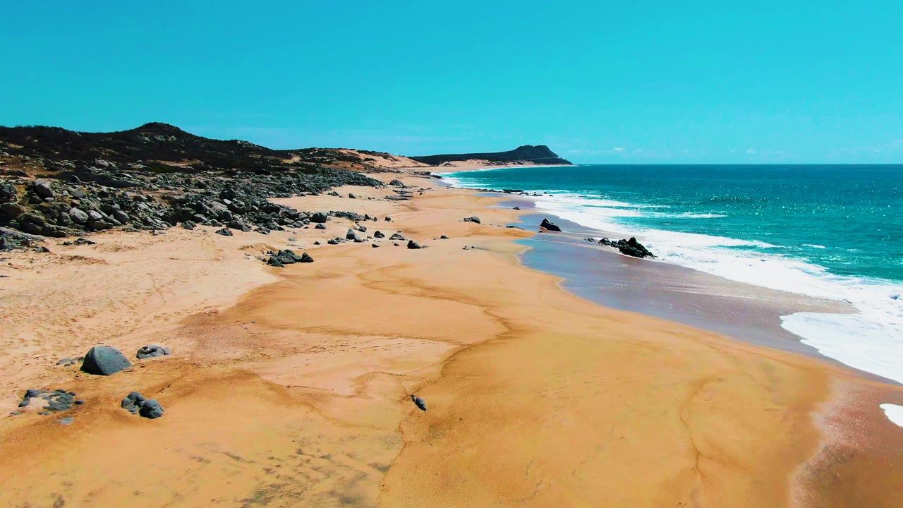 A high aerial view over Buzzards Beach at Punta Buzzards shows golden sand, scattered dark rocks, turquoise surf along the right edge, distant low hills, and no visible people.