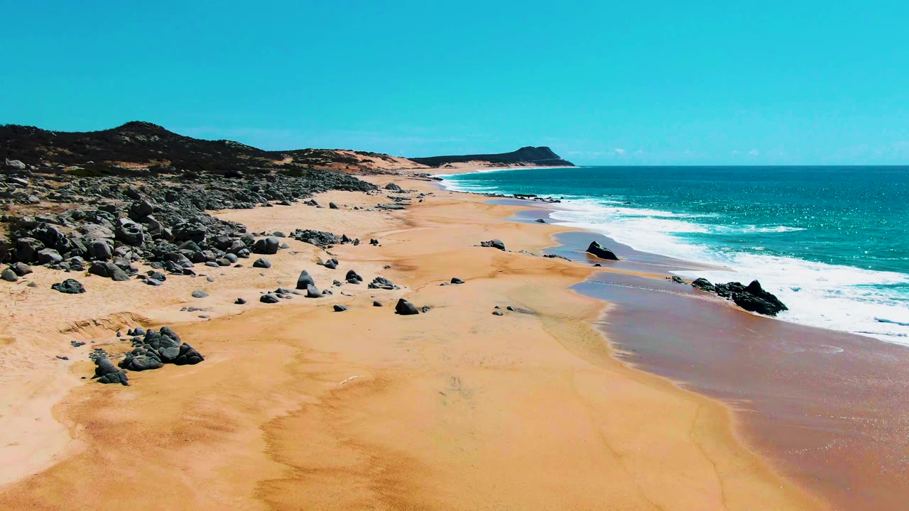 A slightly later view shows more wet sand patterns near the waterline at Buzzards Beach, with dark rocks spread across the left midground, low hills behind, and no visible people.