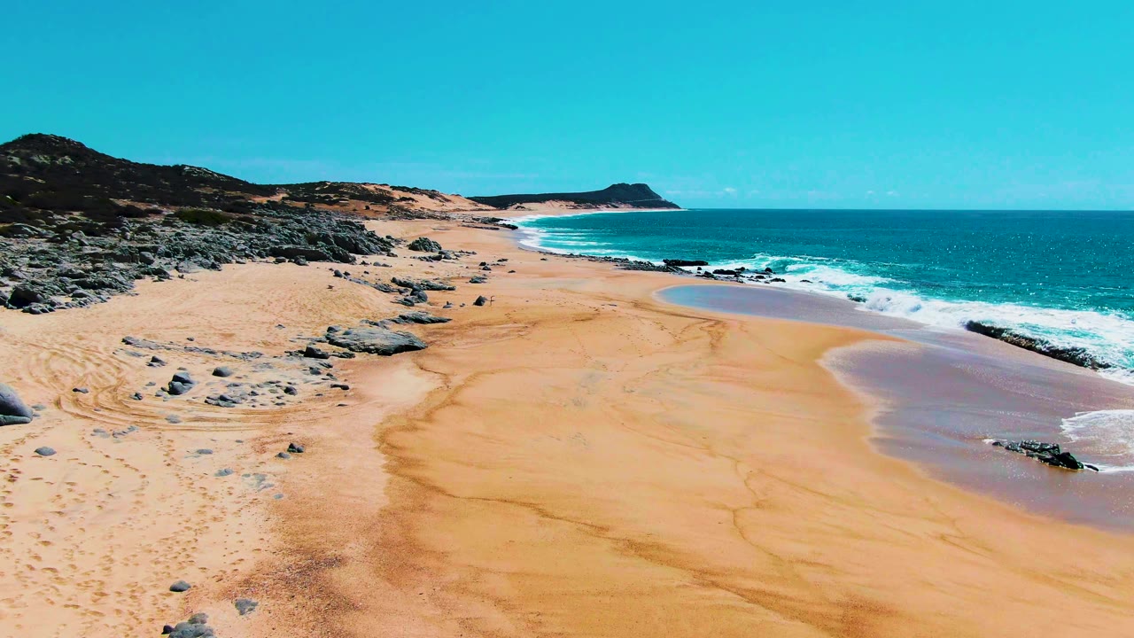 Aerial frame over Buzzards Beach in Baja California Sur showing broad sand, gray rocks at left, white surf on the right, distant low hills, and no visible people.