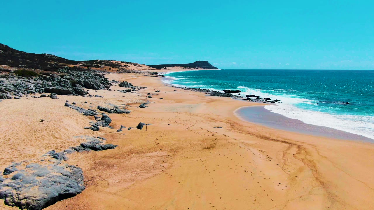 Drone-height view of Buzzards Beach with foreground rocks entering from the left, a wide empty sand flat, breaking foam near the waterline, and no visible people.