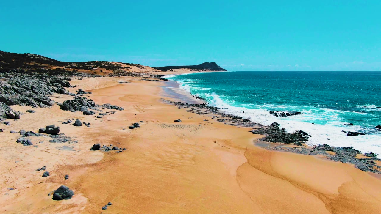 Aerial view over Buzzards Beach in Punta Buzzards, Baja California Sur, with golden sand, dark rocks, turquoise ocean, white surf, low coastal hills, and no visible people.