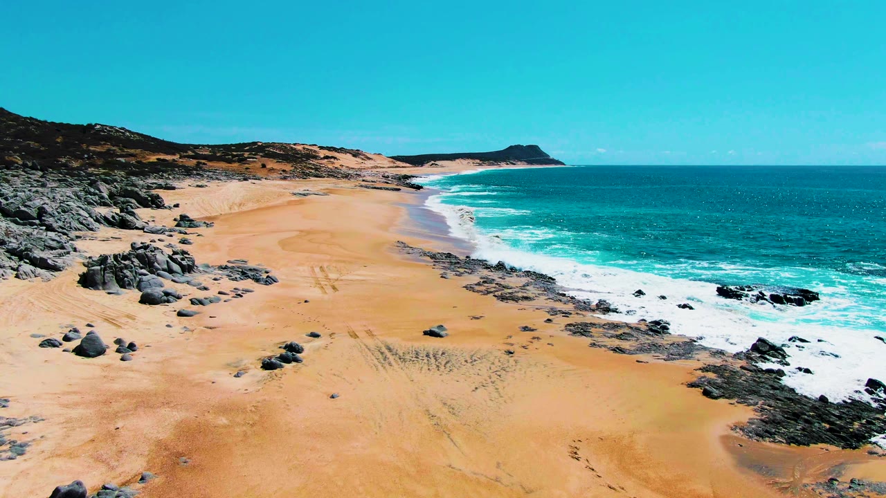 Drone view along Buzzards Beach with more exposed sand in the foreground, scattered rocks on the left, surf breaking along the right edge, and no visible people.