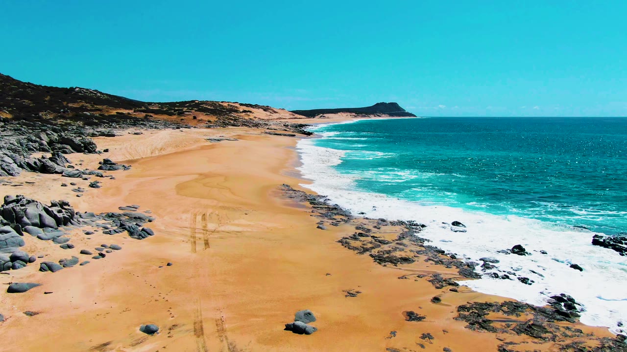 Wide aerial frame at Punta Buzzards showing a broad sandy beach with faint track marks, rocky areas near the waterline, turquoise sea, foaming waves, and no visible people.