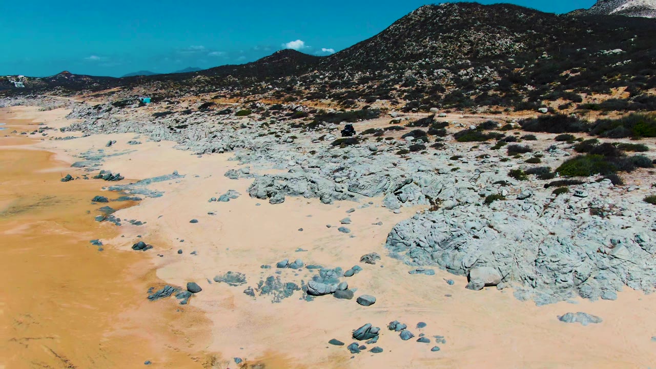 At Punta Buzzards in Baja California Sur, an aerial frame shows pale sand, gray rocks, scrub, a dirt road near the midground, low mountains, blue sky, and no visible people.
