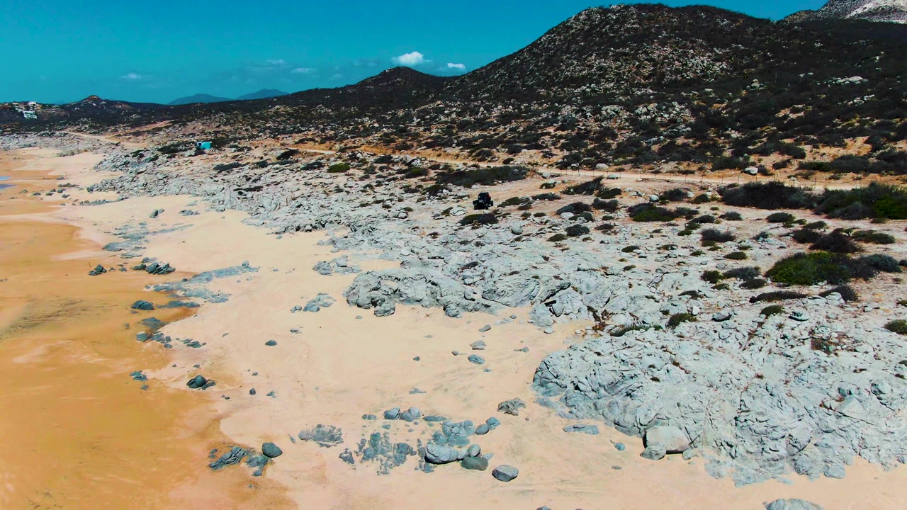 At 0:01, the aerial view holds over sandy ground and pale rock shelves, with the dirt road cutting across the scene below dark shrub-covered hills and no visible people.