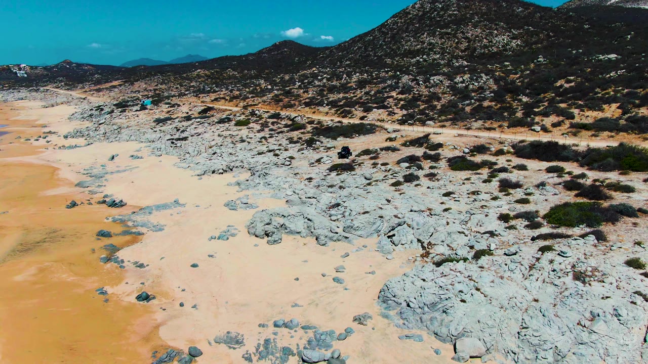 At 0:02, more of the dirt road and scrubby slope fills the right side while pale sand and scattered rocks remain visible on the left, with no visible people.