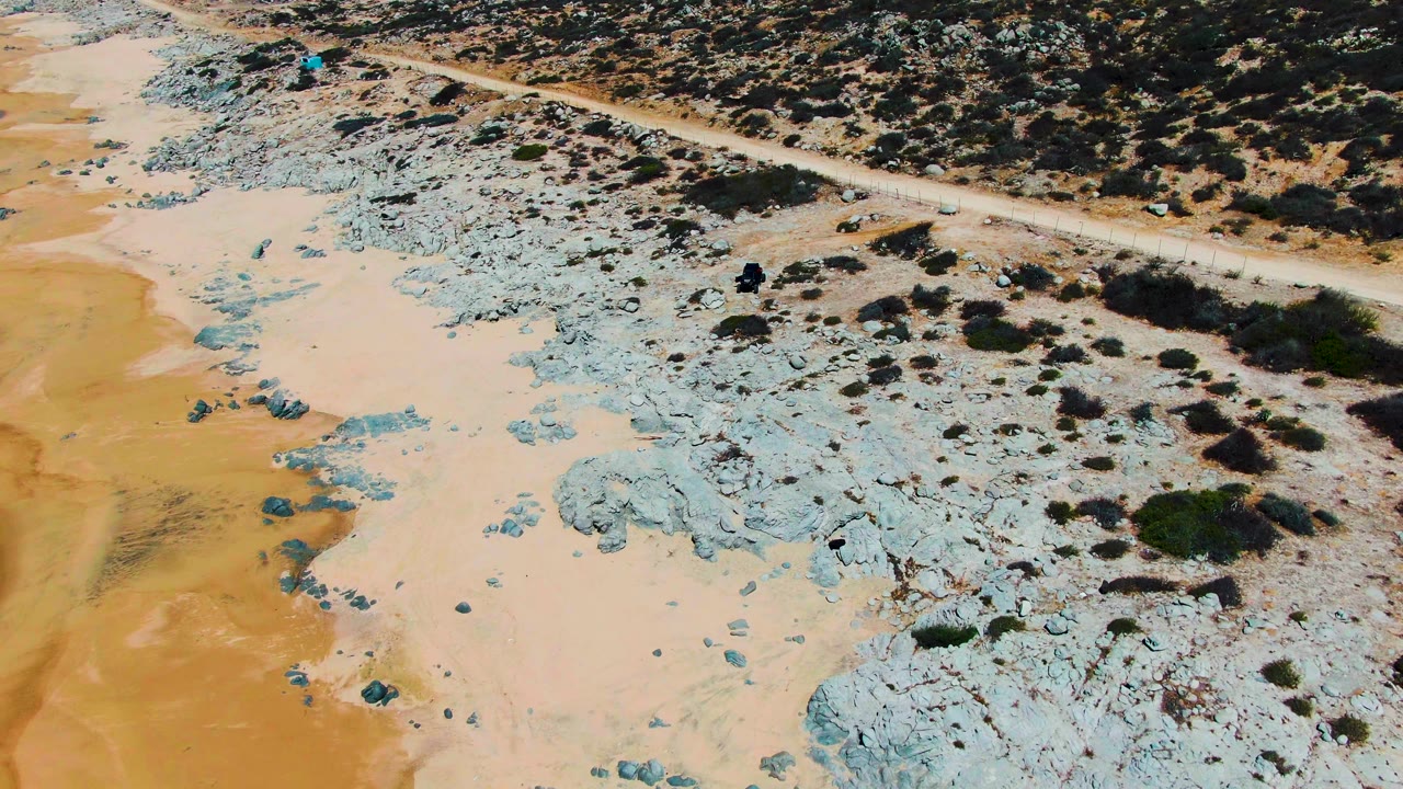 Aerial frame over Punta Buzzards showing a dirt road above blue-gray rock, beige sand, and scattered desert bushes with no visible people.