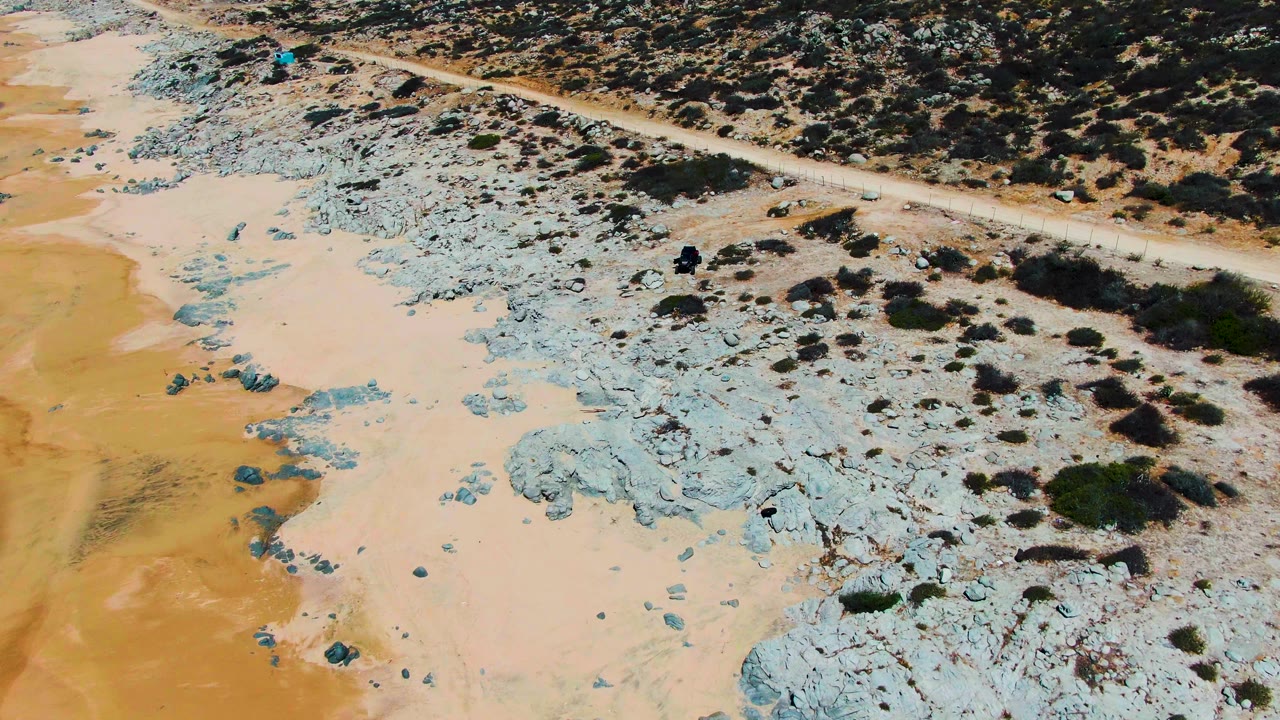 High aerial frame at Punta Buzzards with the dirt road running across the top of blue-gray rock formations and pale beach sand, no visible people.
