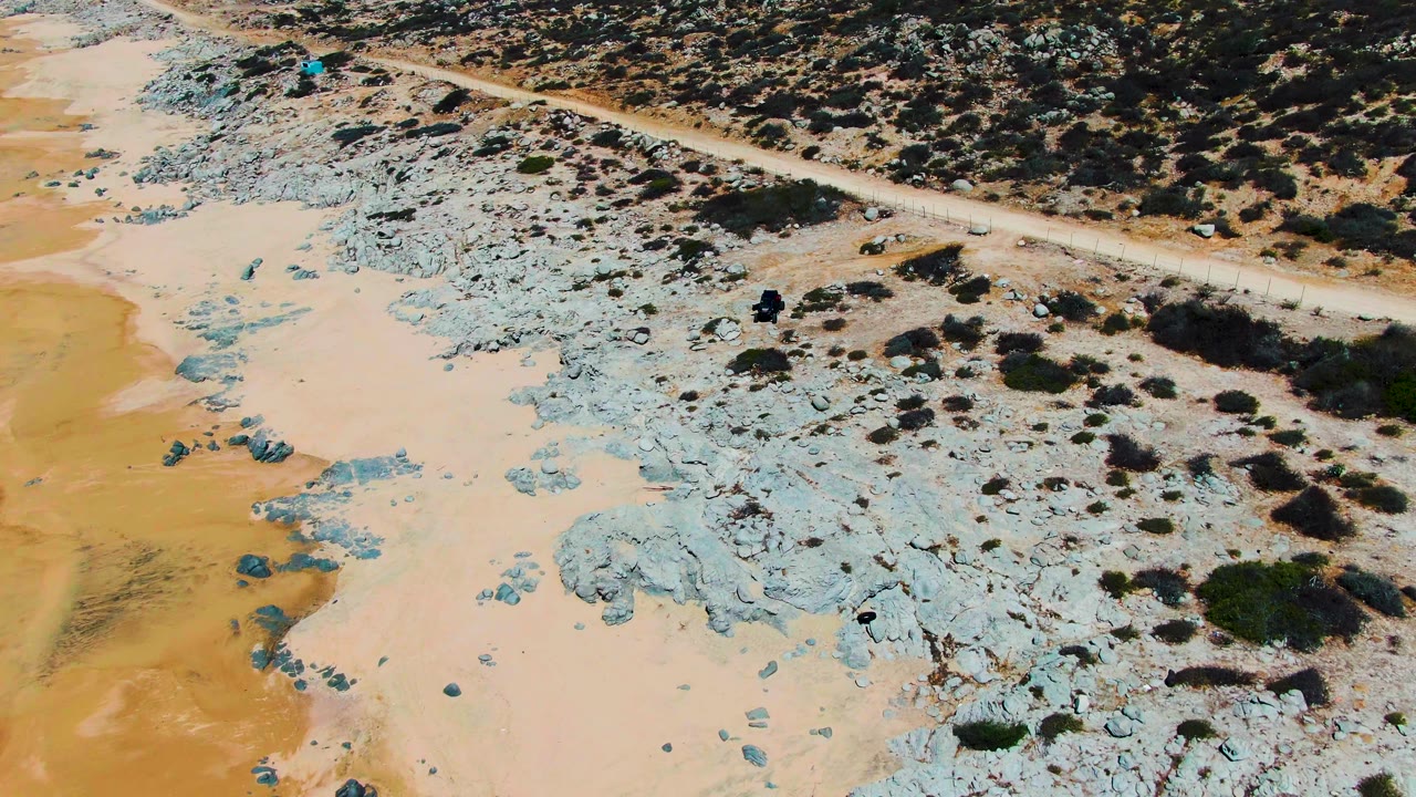 Overhead Punta Buzzards view with scrub-covered brown ground, a pale dirt road, and blue-gray rocks meeting beige sand, with no visible people.