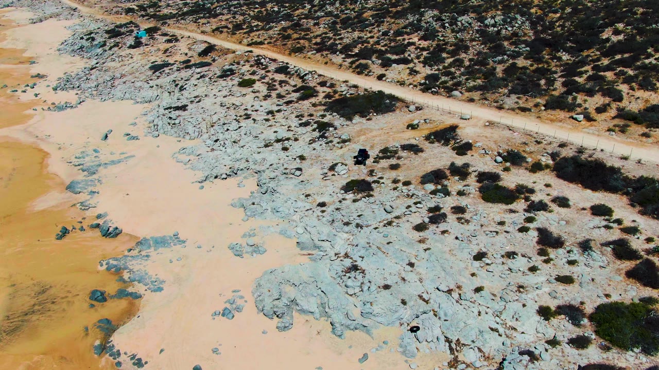 Aerial view closer to the beach edge with rocks, sand, bushes, and road.