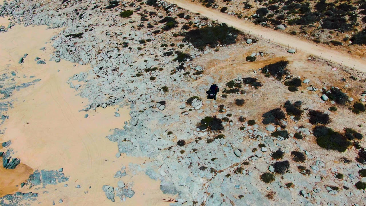 Overhead frame from Punta Buzzards, Baja California Sur, showing a dark vehicle near pale rock, tan sand, dirt tracks, and scattered shrubs with no visible people.