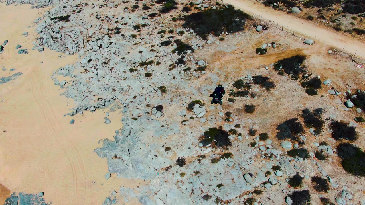 Aerial view in Punta Buzzards, Baja California Sur, with the dark vehicle slightly lower among pale rocks, small boulders, scrub patches, and a sandy path with no visible people.