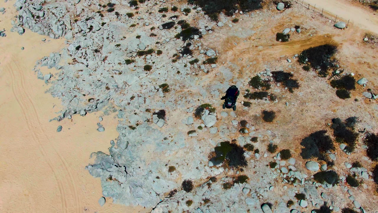 Overhead Punta Buzzards frame showing the dark vehicle farther into the rocky area, with tan dirt, pale stone, isolated shrubs, and no visible people.