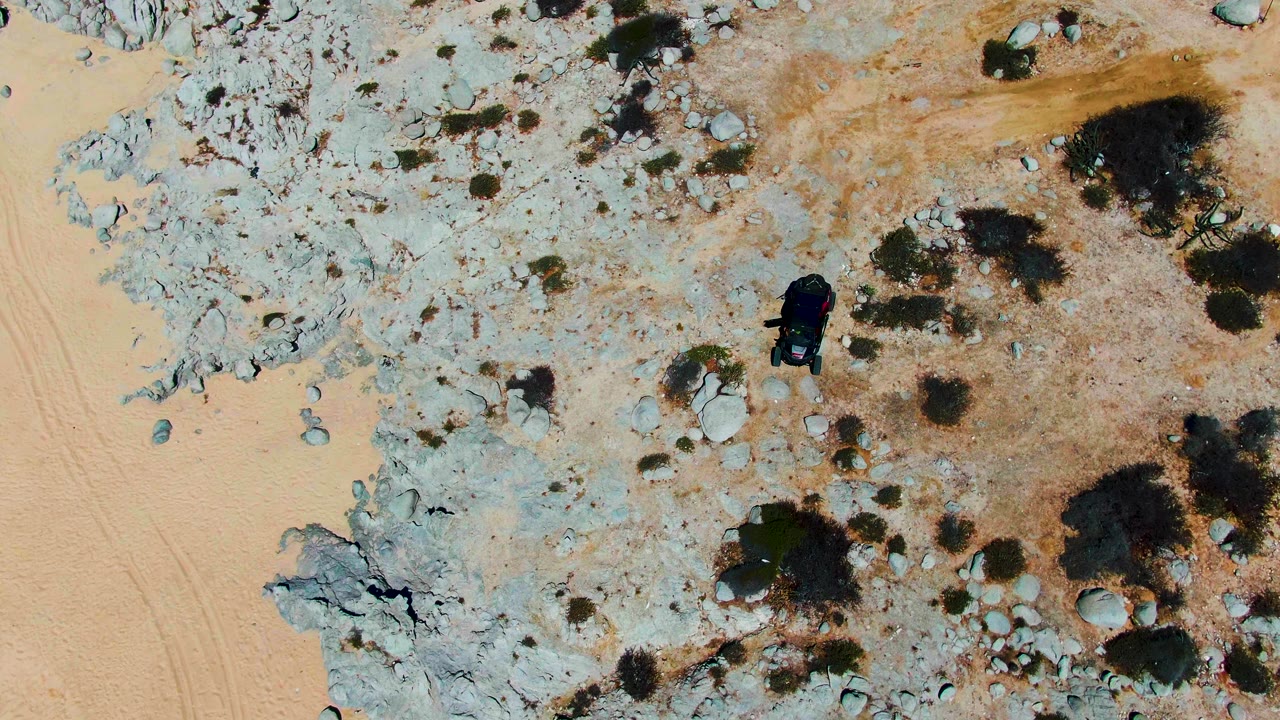 Top-down view at Punta Buzzards with the vehicle centered lower than before, surrounded by pale rock slabs, scattered boulders, dry shrubs, and tan sand with no visible people.
