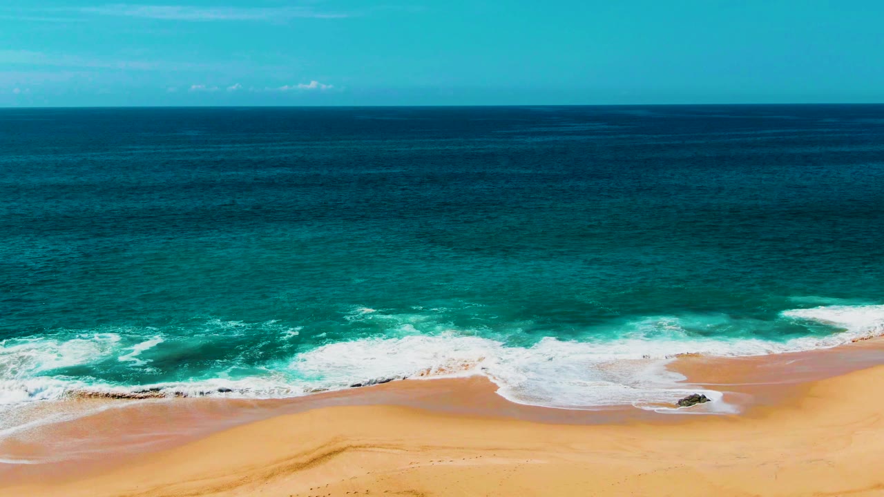 A second later, the white foam stretches farther across the shore at Punta Buzzards, with dark blue water beyond, golden sand below, and no visible people.