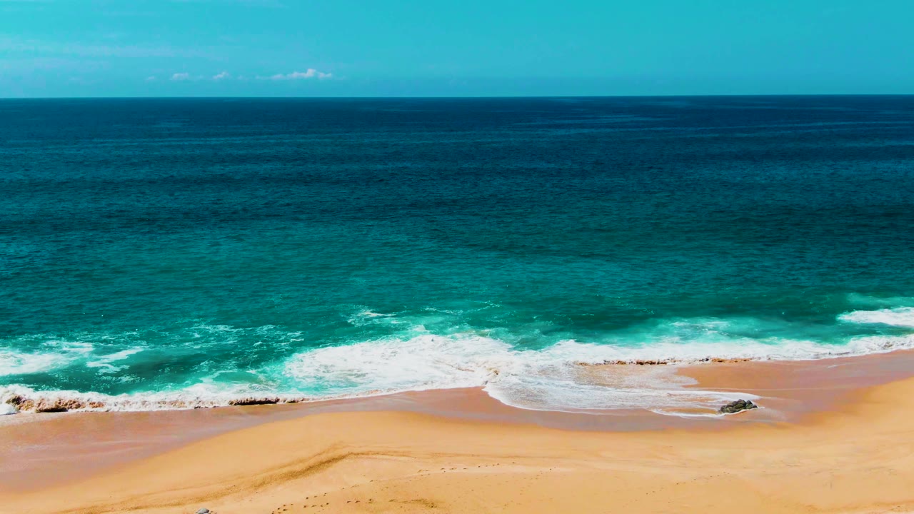 The surf line at Punta Buzzards appears wider across the frame, with foam washing toward smooth sand, scattered rocks near the water, and no visible people.