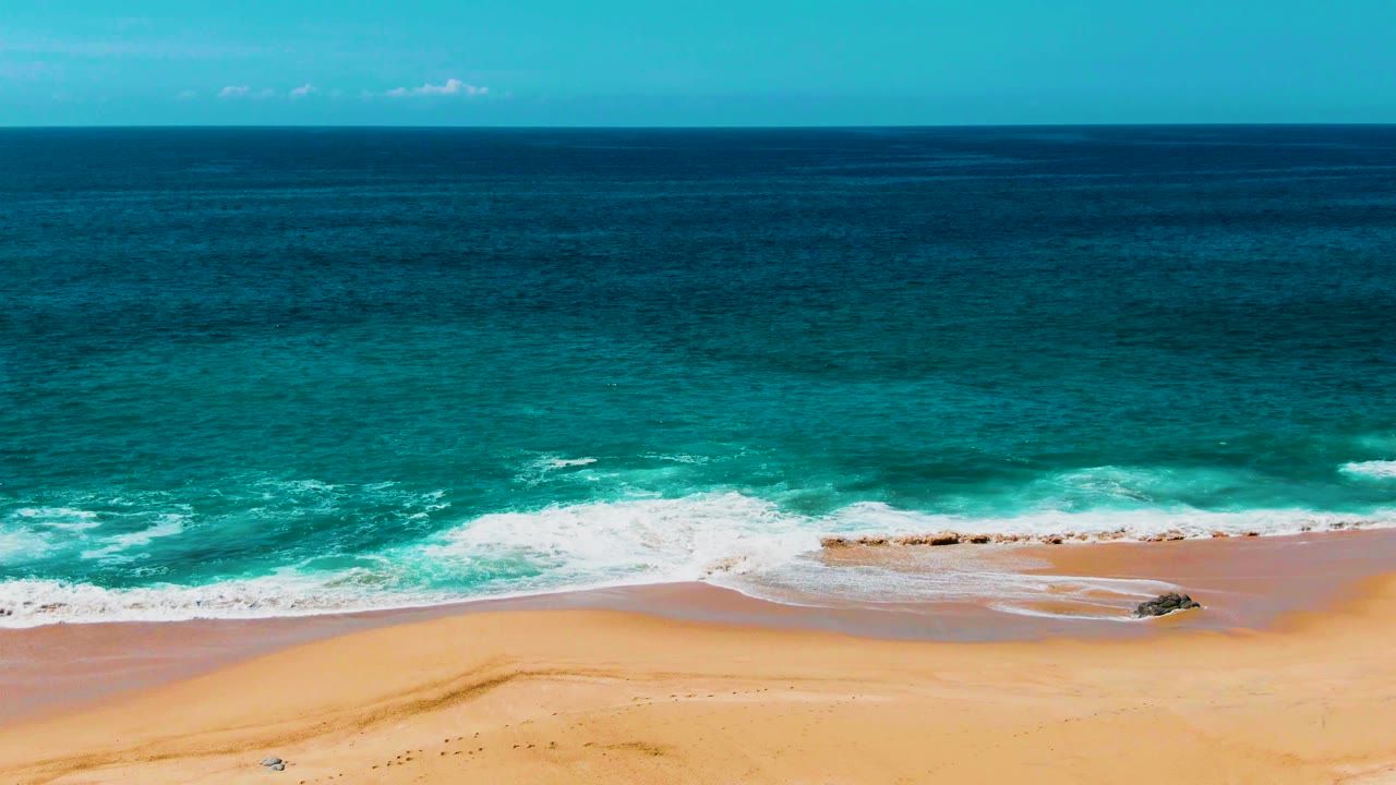 At 0:03, the aerial view holds on Punta Buzzards as foam spreads over wet sand, darker ocean water sits beyond the turquoise band, and no visible people appear.