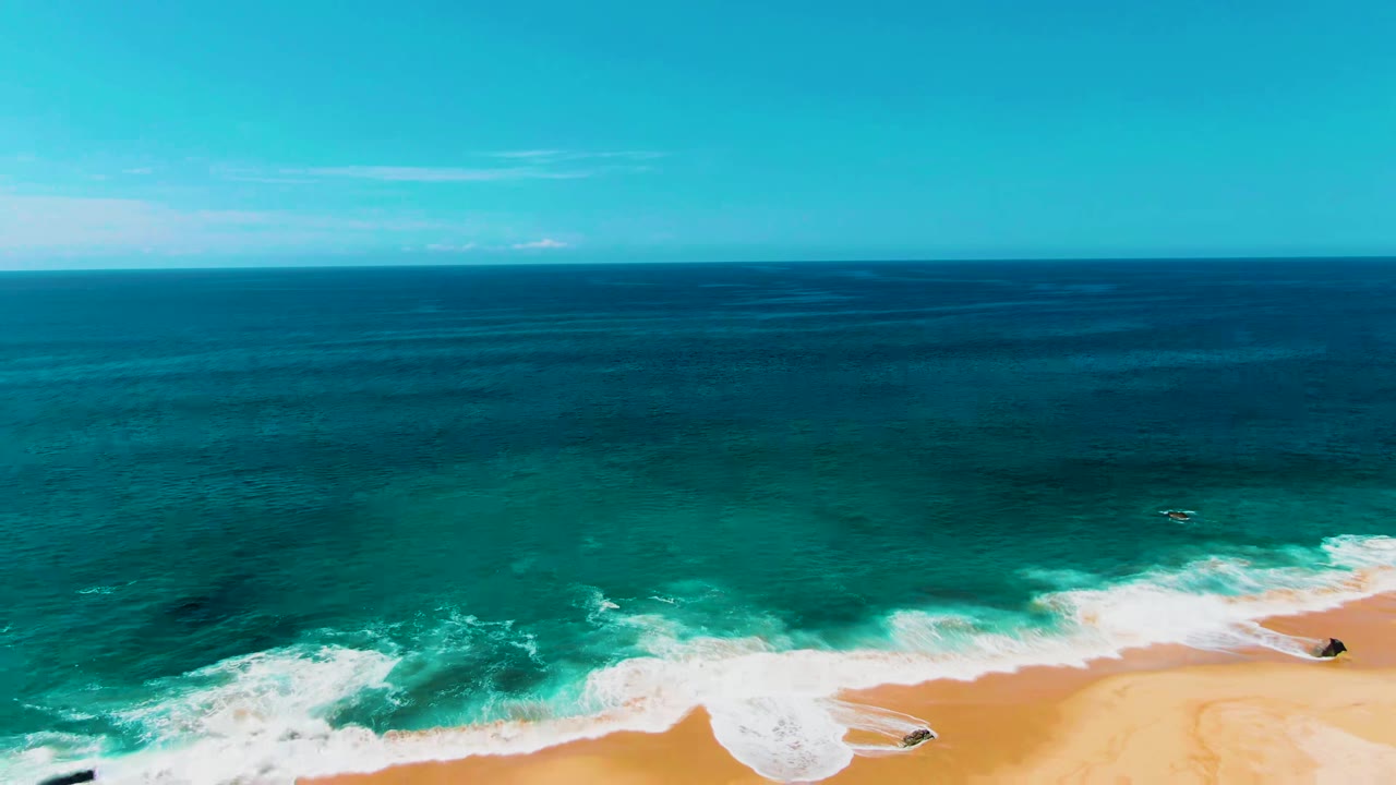 Opening frame over Punta Buzzards, Baja California Sur, with deep blue ocean, turquoise surf, golden sand, small dark shoreline rocks, and no visible people.