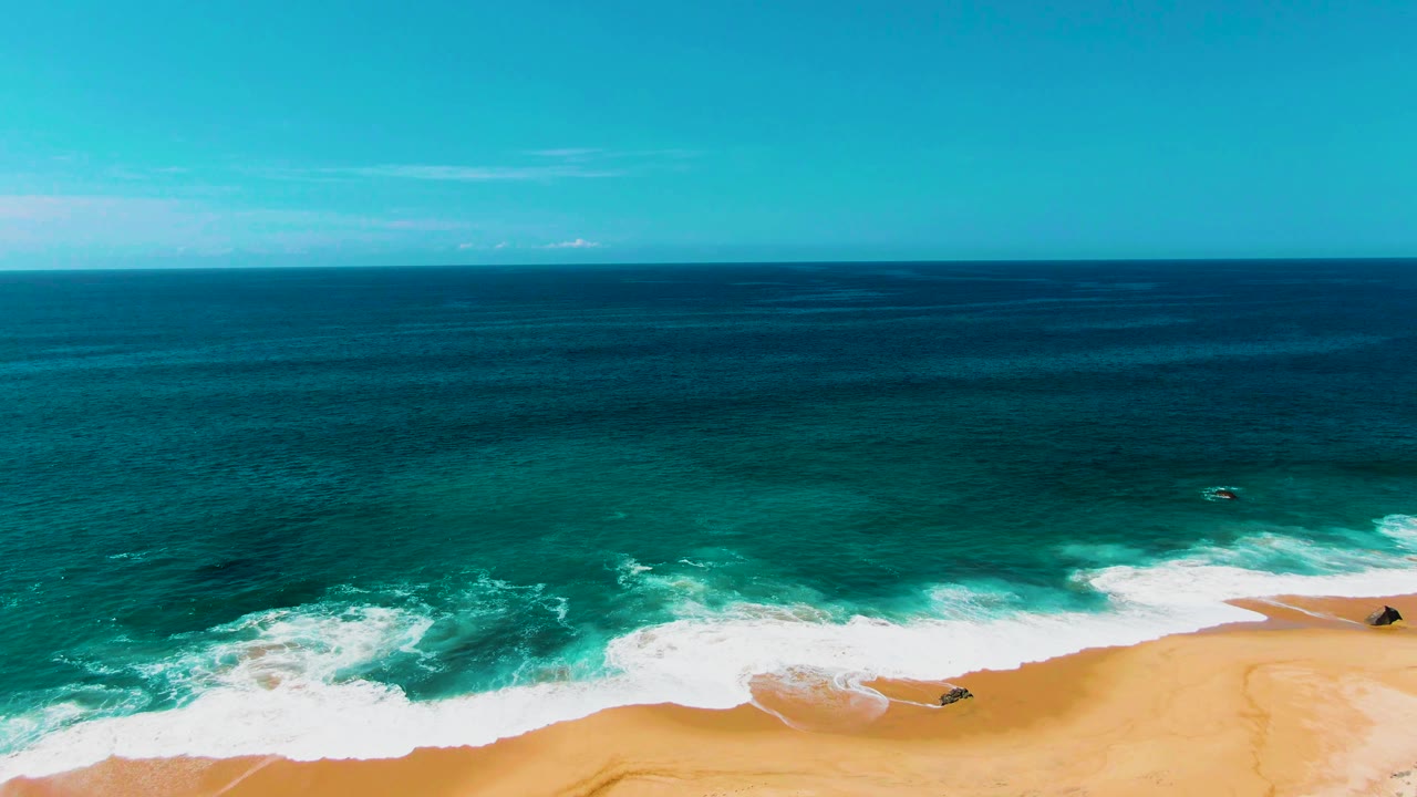 At 0:02, the Punta Buzzards shoreline curves through the lower frame as broken white water spreads over golden sand, with no visible people.