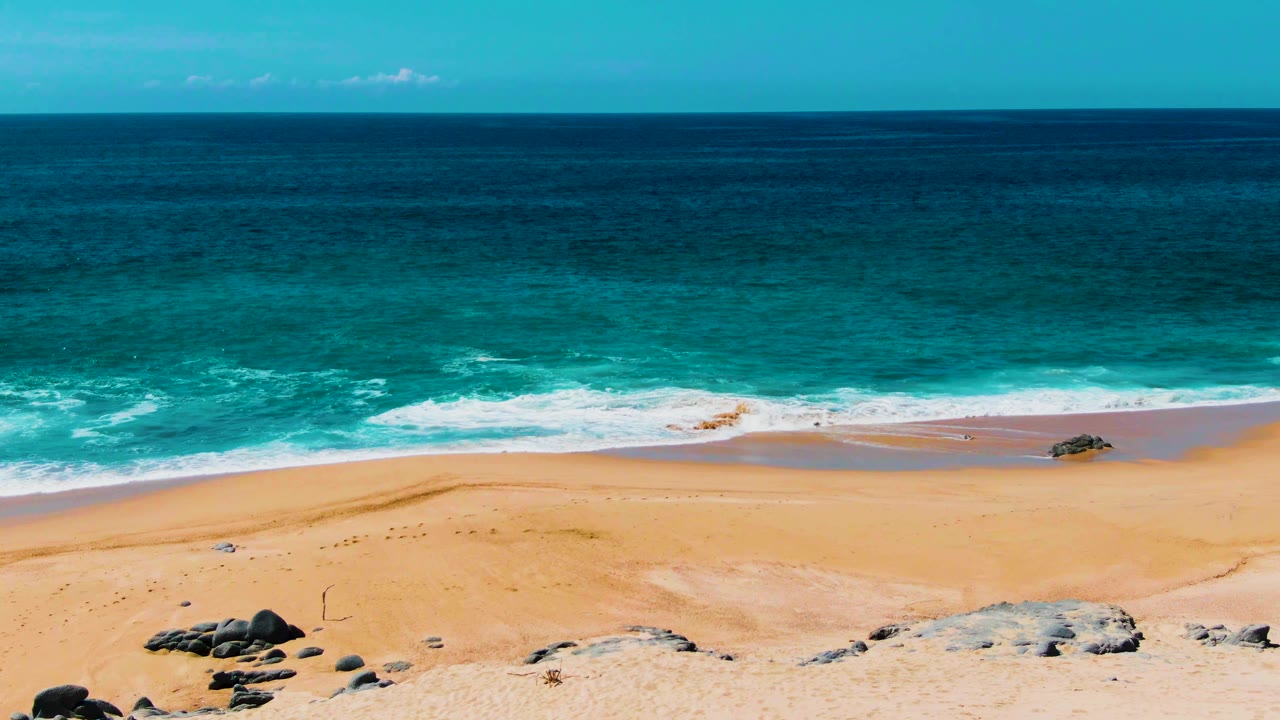Wide view of Buzzards Beach in Punta Buzzards, Baja California Sur, with turquoise ocean, white surf, tan sand, scattered rocks, and no visible people.
