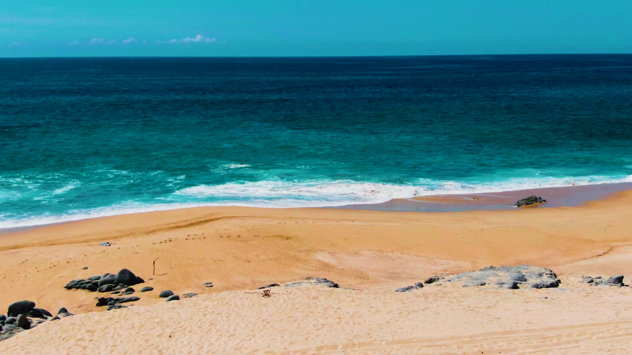 Buzzards Beach shoreline in Baja California Sur shows a darker band of ocean, white wave foam, open sand, rock clusters, and no visible people.