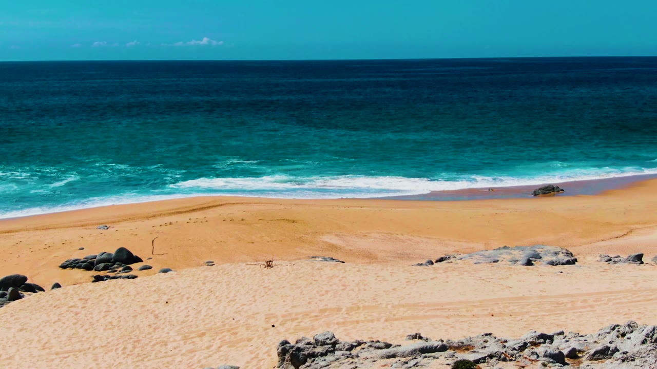The Punta Buzzards beach view holds on turquoise water, a white surf line, broad sand, foreground rock formations, and no visible people.