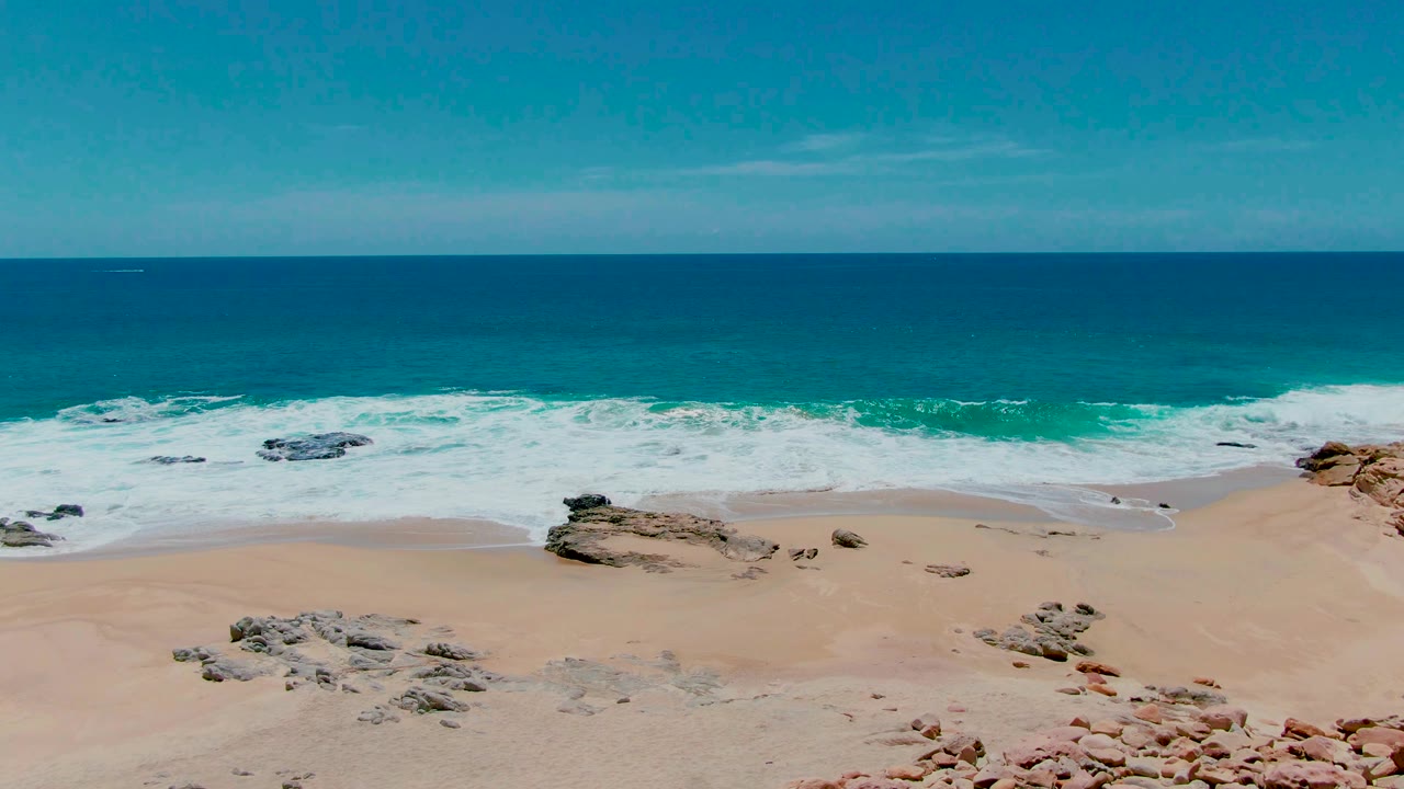 Aerial view of sandy beach, rocks, turquoise surf, and deep blue ocean.