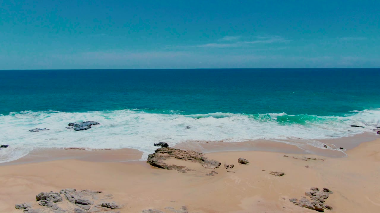 Aerial frame at Punta Buzzards, Baja California Sur with foamy surf advancing around exposed rocks on pale sand, deep blue ocean behind, and no visible people.