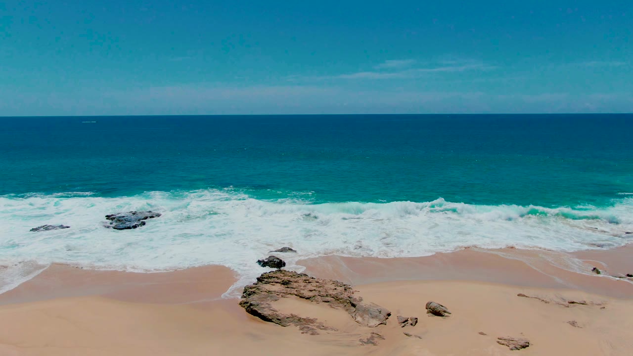 Aerial frame at Punta Buzzards, Baja California Sur showing a larger rock formation near the lower center, foamy surf behind it, open ocean, and no visible people.