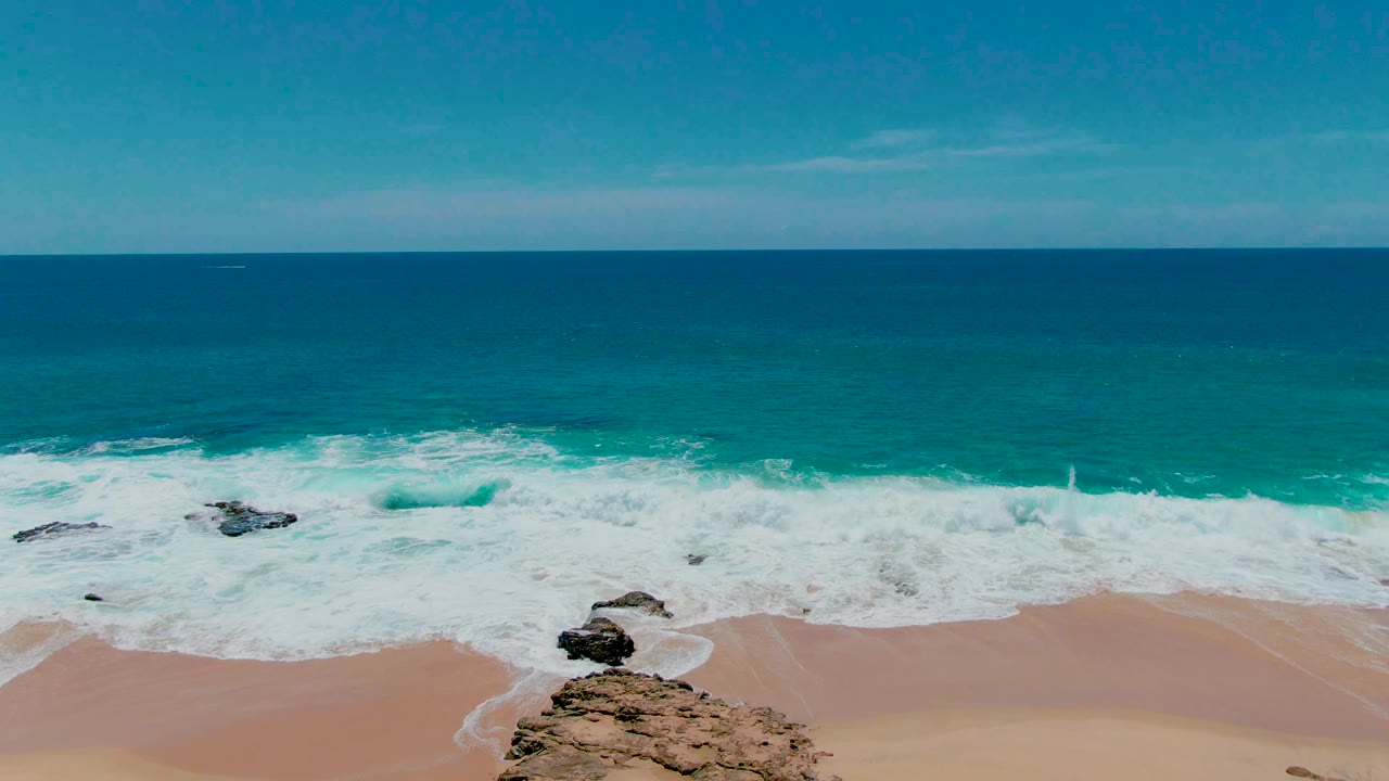Aerial frame at Punta Buzzards, Baja California Sur with less visible sand, surf filling more of the frame, small rocks near the shoreline, and no visible people.