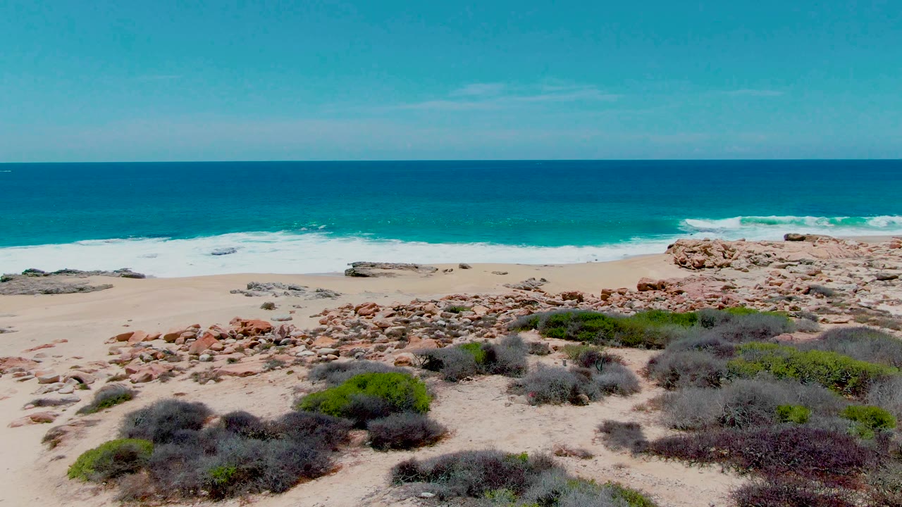 Buzzards Beach seen from above with less foreground vegetation, more exposed sand and rocks, white waves rolling toward shore, blue ocean, and no visible people.