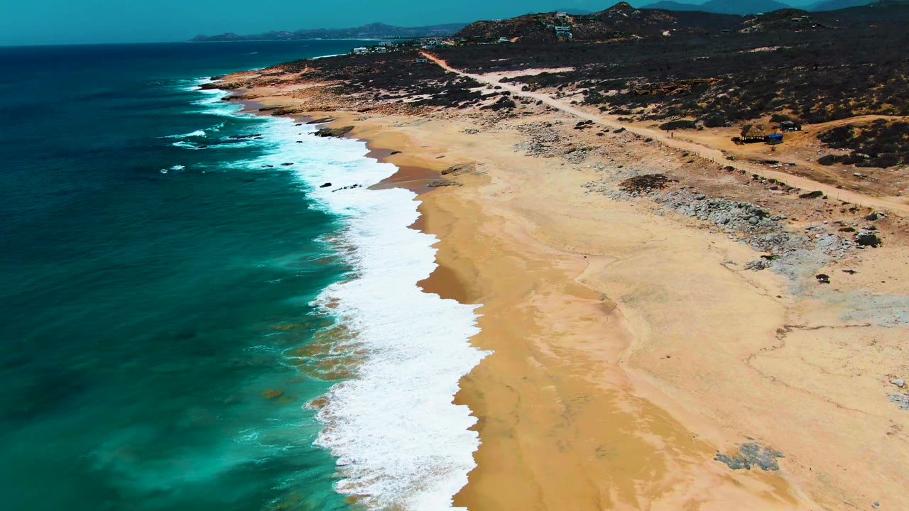 Oblique aerial view over Buzzards Beach in Baja California Sur, with turquoise water and white surf entering from the left, broad sand to the right, a dirt coastal road above the beach, and no visible people.